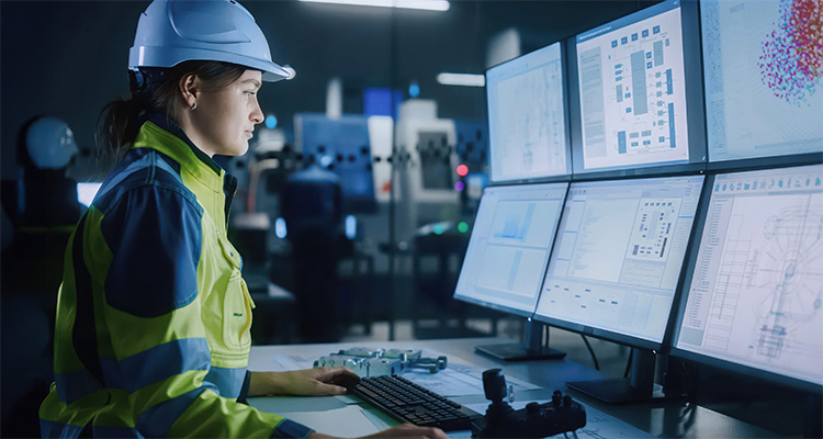 an individual in a hard hat and high-visibility clothing, working in a control room or industrial setting with multiple monitors displaying technical data and schematics.