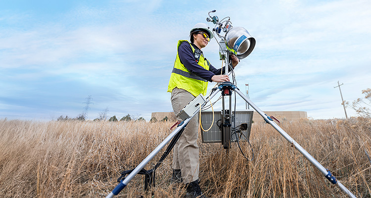 Montrose Environmental Group tackles the planet’s toughest challenges by turning emissions data into actionable intelligence a person wearing personal protective equipment, including a white hard hat, safety glasses, and a high-visibility yellow vest, operating a piece of equipment in a field of tall, dry grass