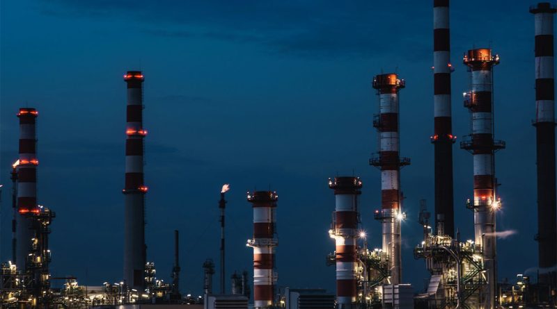 Oil refinery at night with illuminated industrial towers and red warning lights on tall smokestacks.